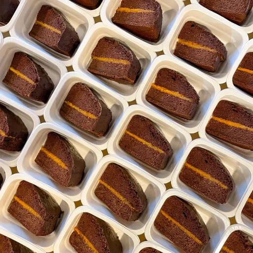 Multiple slices of chocolate cake with orange filling neatly arranged in white plastic containers.