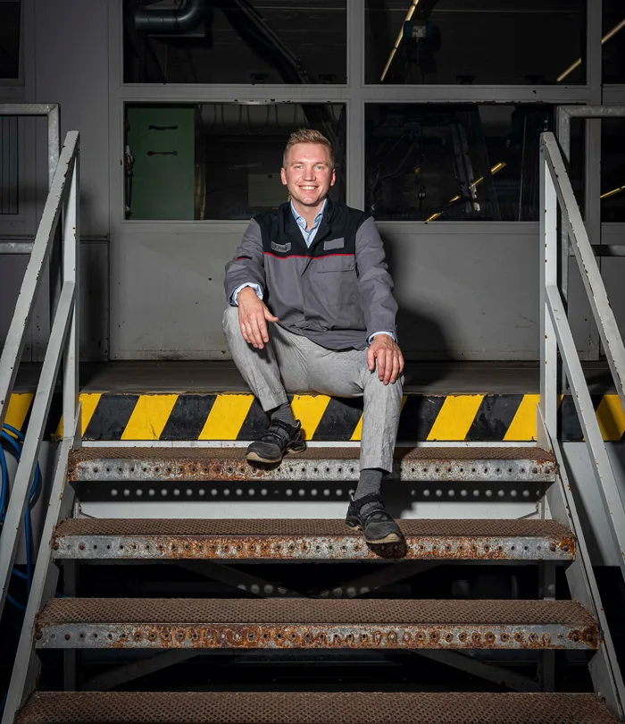 Junger Mann in grauer Arbeitsjacke sitzt auf einer Metalltreppe in einem industriellen Umfeld und lächelt in die Kamera.