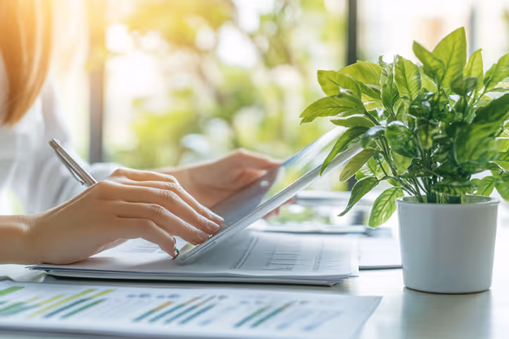 Nahaufnahme einer Hand mit Tablet auf einem Schreibtisch mit Auswertungen und grüner Zimmerpflanze – digitaler, papierarmer Versand als umweltfreundliche Alternative.