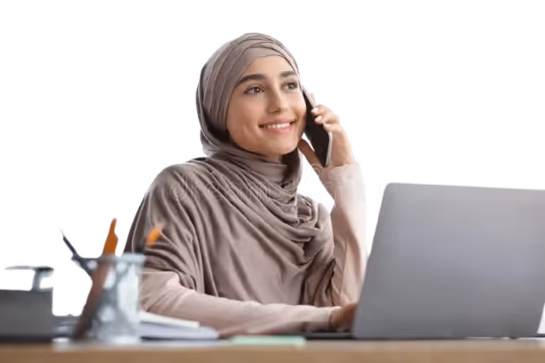 Smiling woman wearing a hijab talking on a smartphone while using a laptop at a desk.