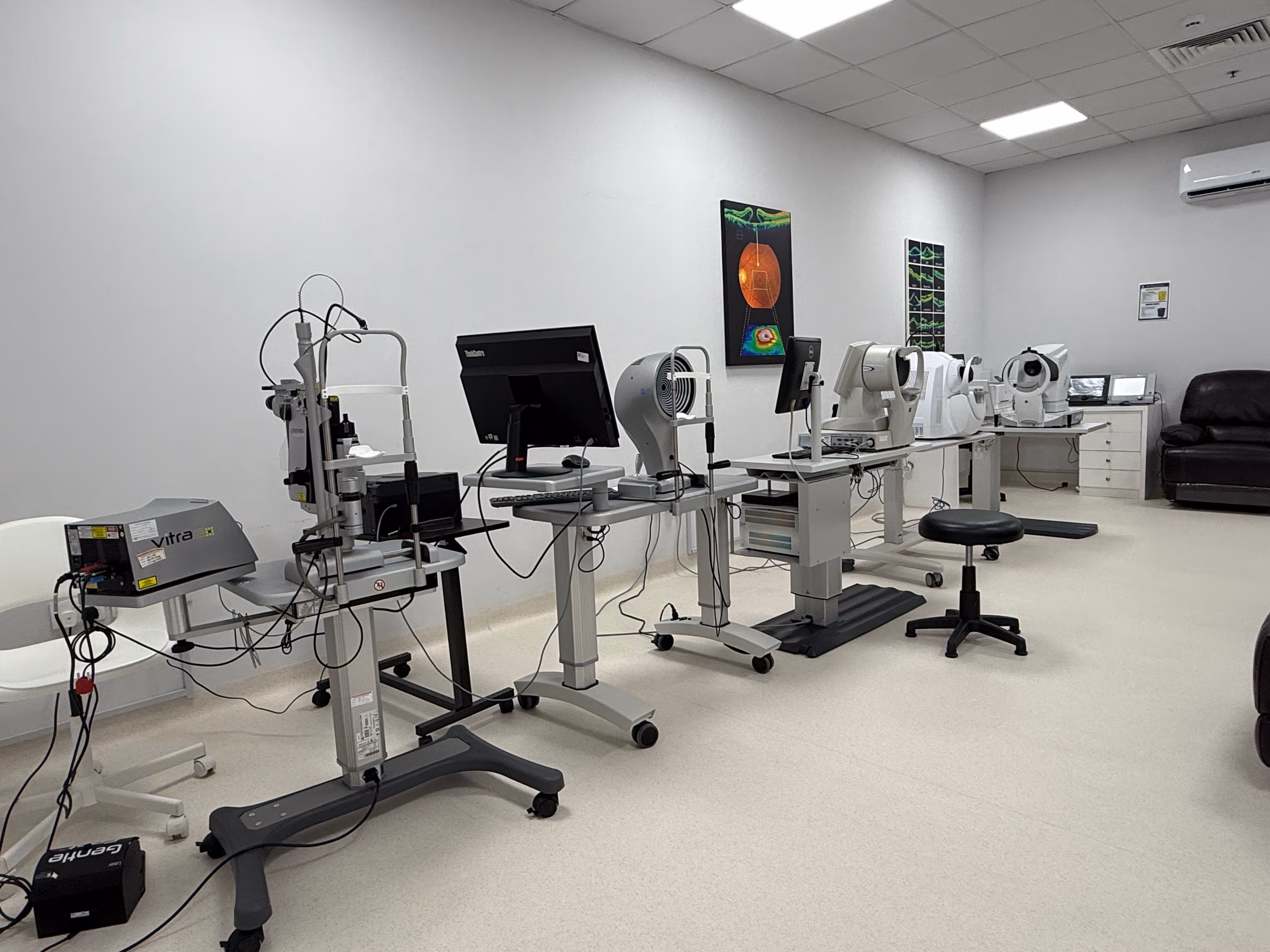 A clinical room with several ophthalmic diagnostic machines lined up against a white wall, accompanied by chairs and computers.