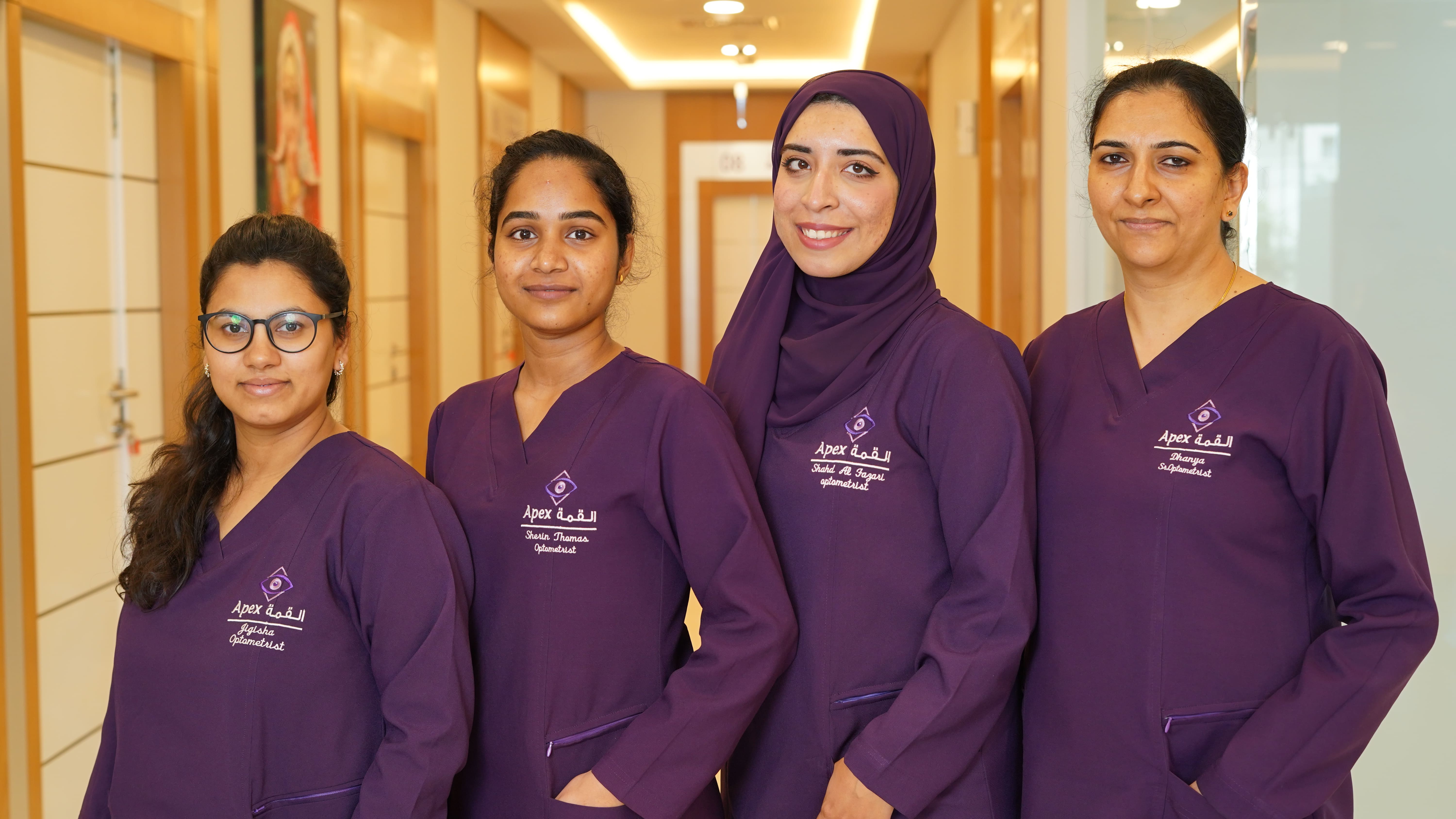 Four female optometrists in purple uniforms standing in a hallway at Apex clinic.