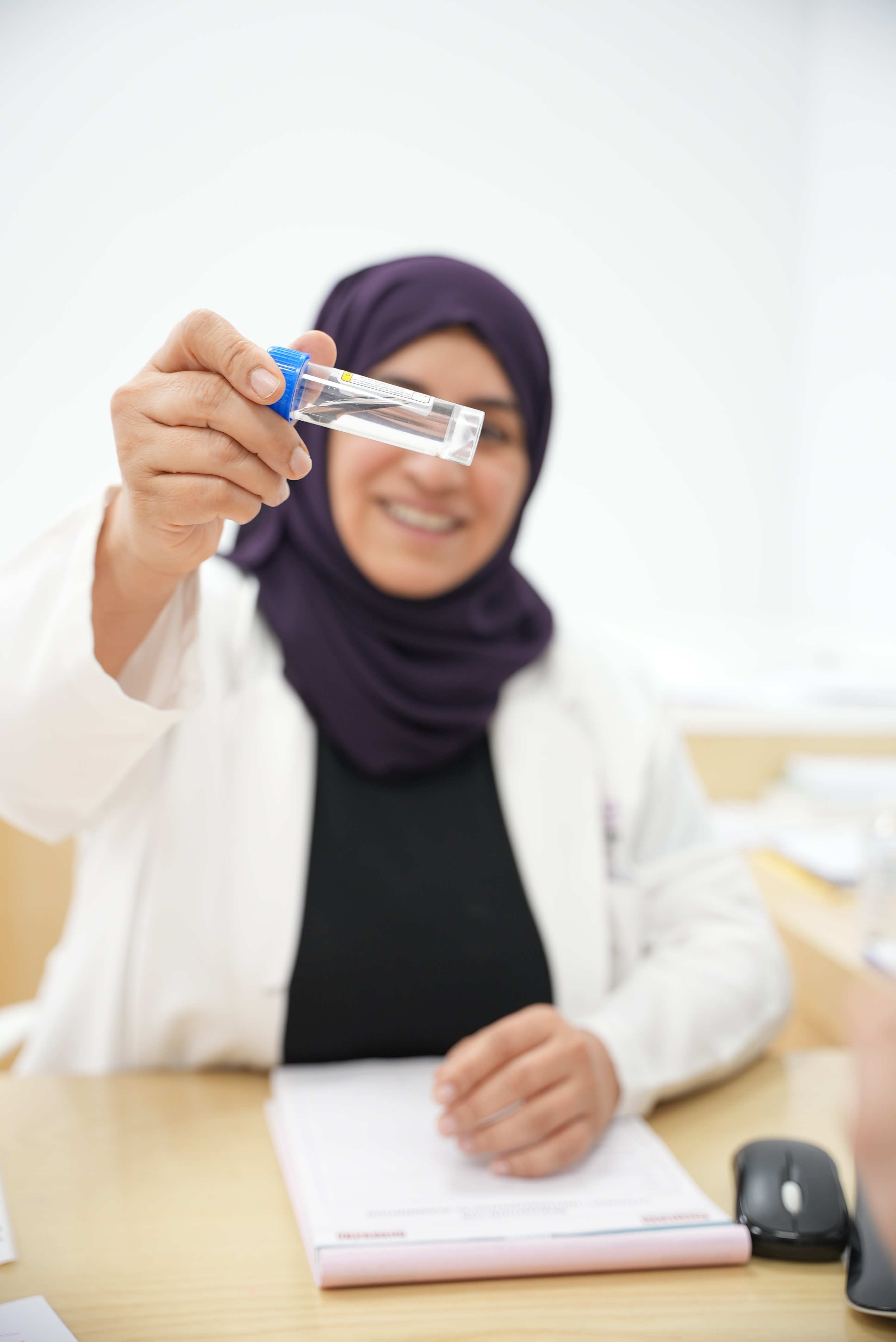 Smiling woman in a white lab coat and dark hijab holding a clear test tube with a blue cap towards the camera.