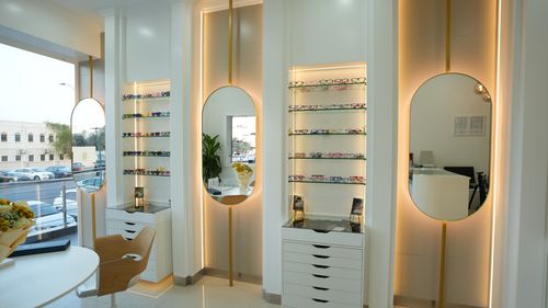 Modern eyewear store interior with illuminated oval mirrors, shelves displaying colorful eyeglasses, and a wooden chair near a white table.