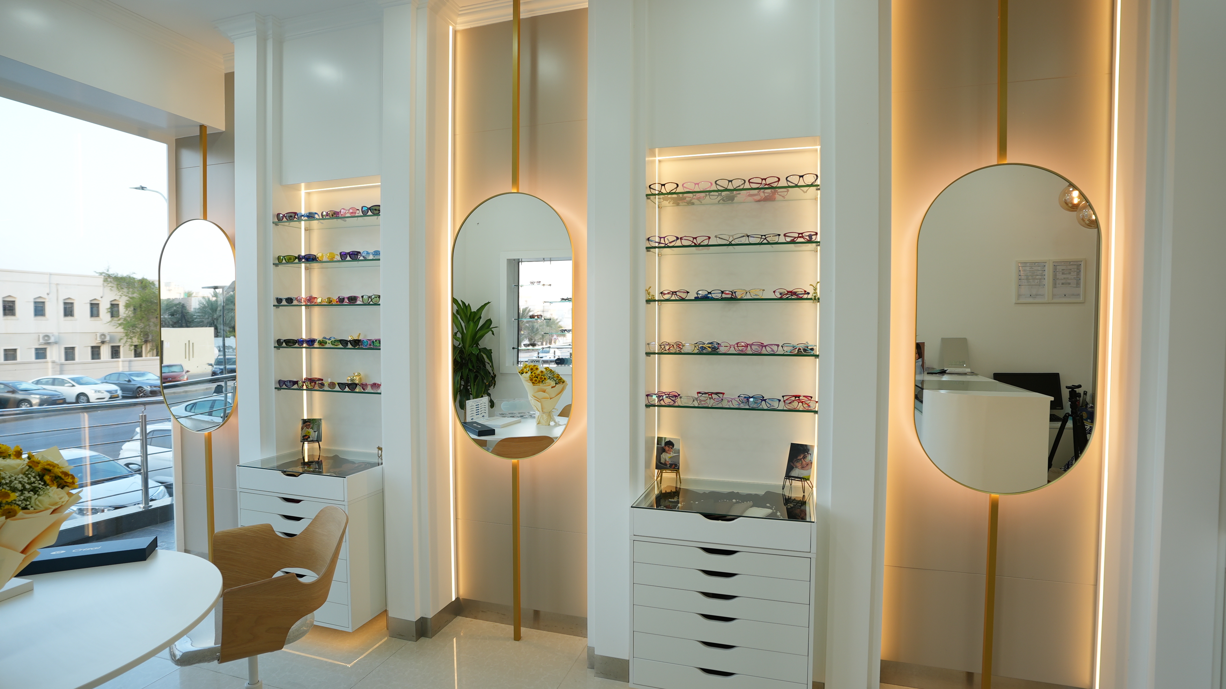 Modern eyewear store interior with illuminated oval mirrors, shelves displaying colorful eyeglasses, and a wooden chair near a white table.