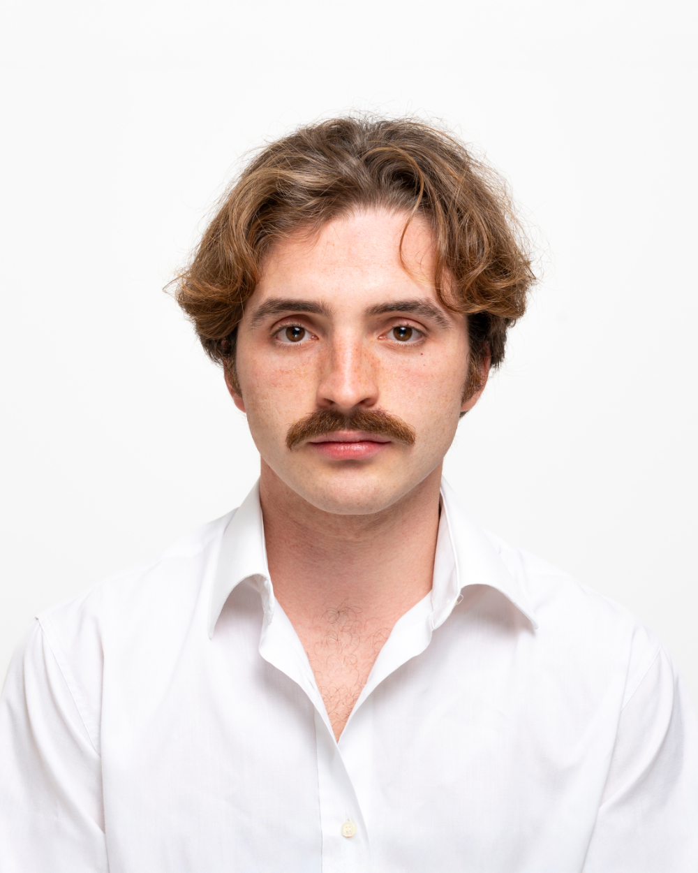 Primo piano di un uomo con baffi castani e capelli ondulati, indossa una camicia bianca su sfondo bianco.