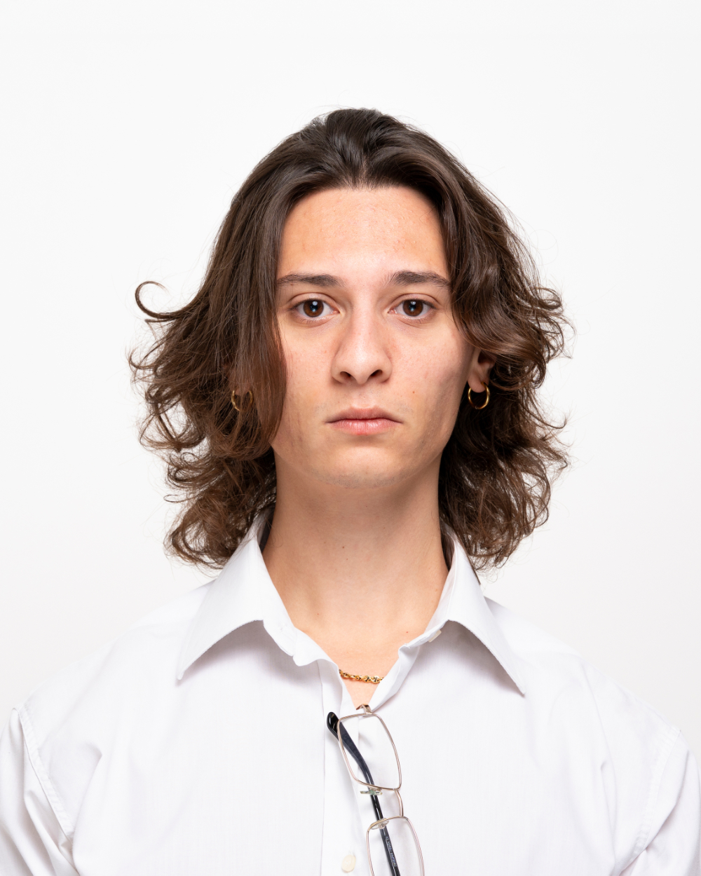 Ritratto frontale di un giovane con capelli castani ondulati, indossa una camicia bianca e orecchini a cerchio dorati.