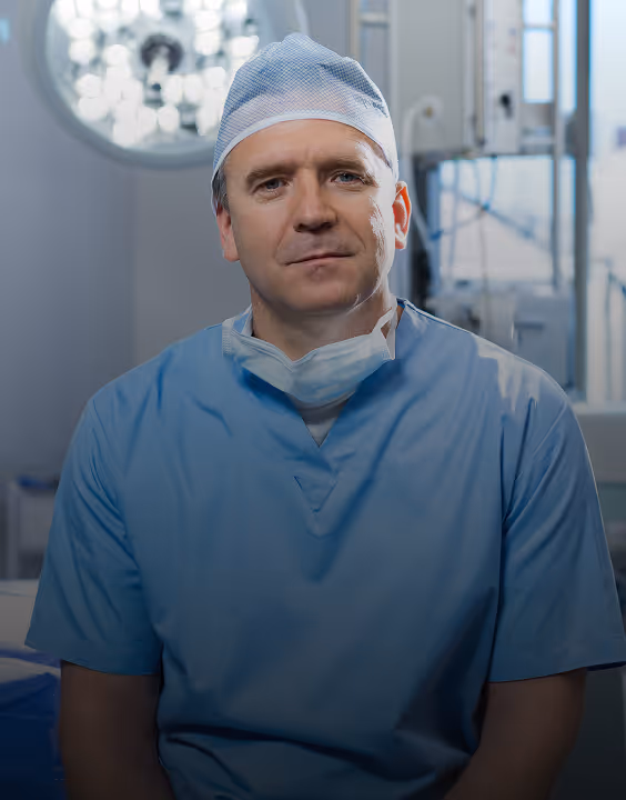 Male surgeon in blue scrubs and surgical cap sitting in an operating room.