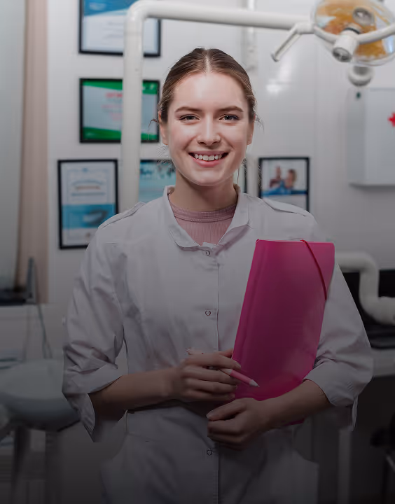 Smiling female dentist in white coat holding a pink folder in a dental clinic.