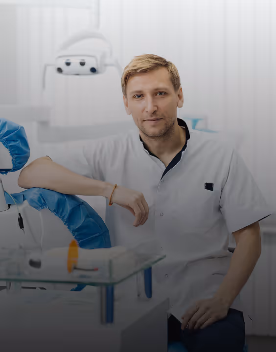 Male dentist in white coat sitting in a dental office with dental equipment in the background.