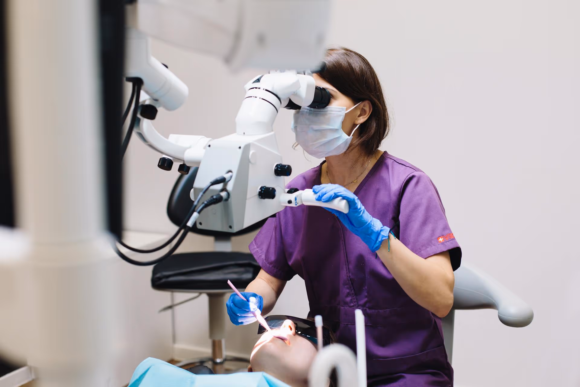 Dentist wearing a purple uniform and mask using a dental microscope while examining a patient’s mouth.