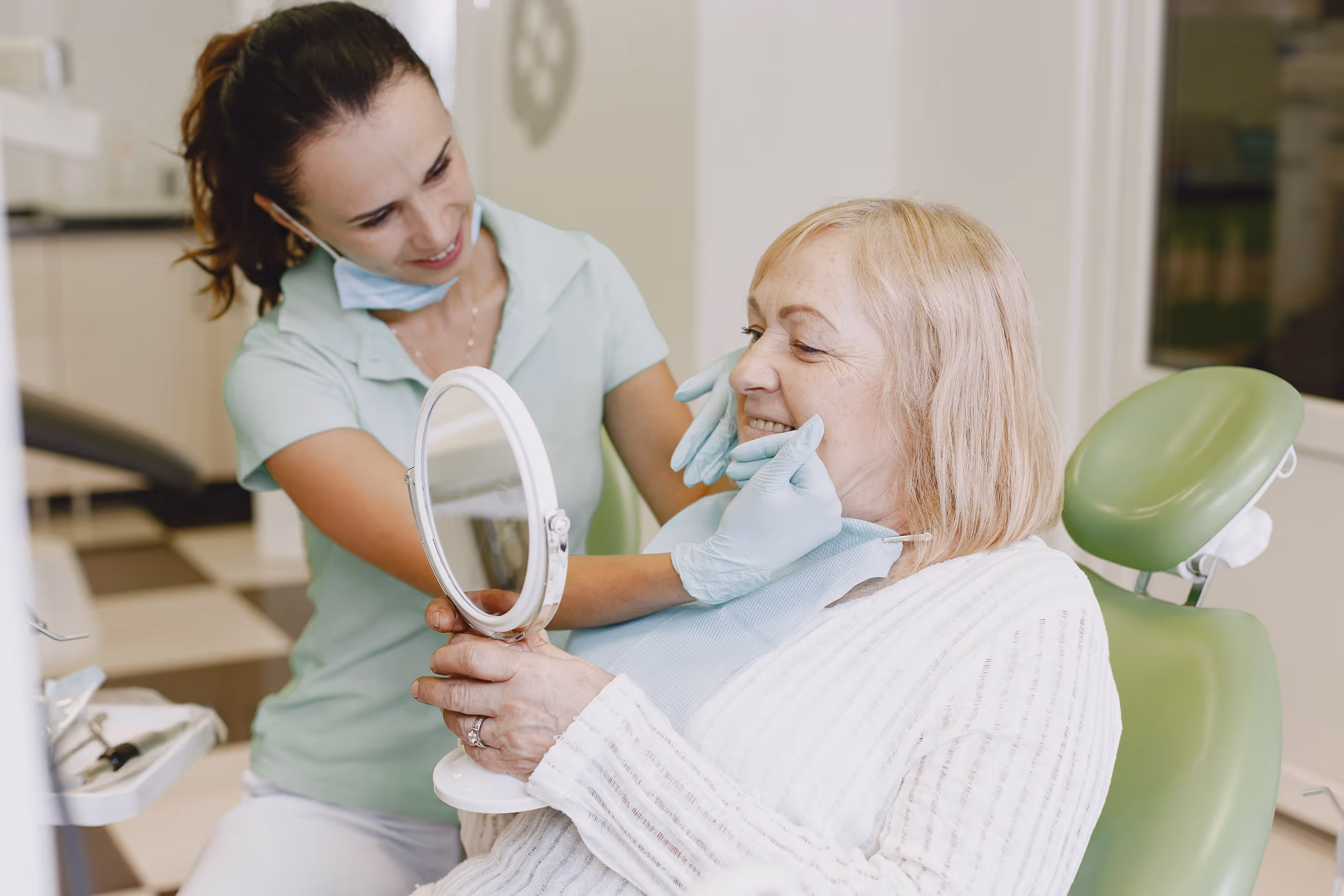 Smiling female dentist in gloves examines an elderly woman's smile while she holds a hand mirror in a dental office.