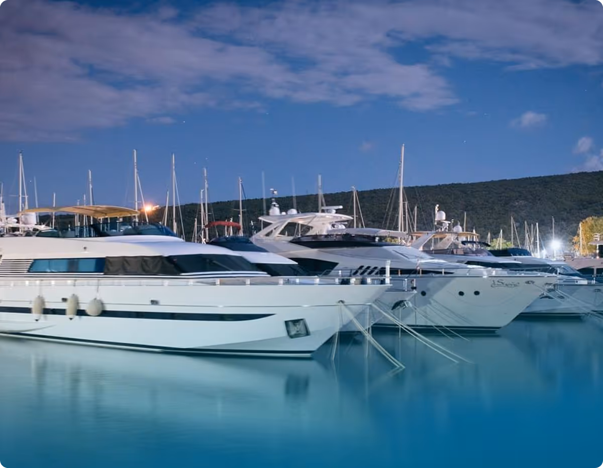 Several luxury yachts docked side by side at a marina during twilight with a partly cloudy sky and forested hills in the background.