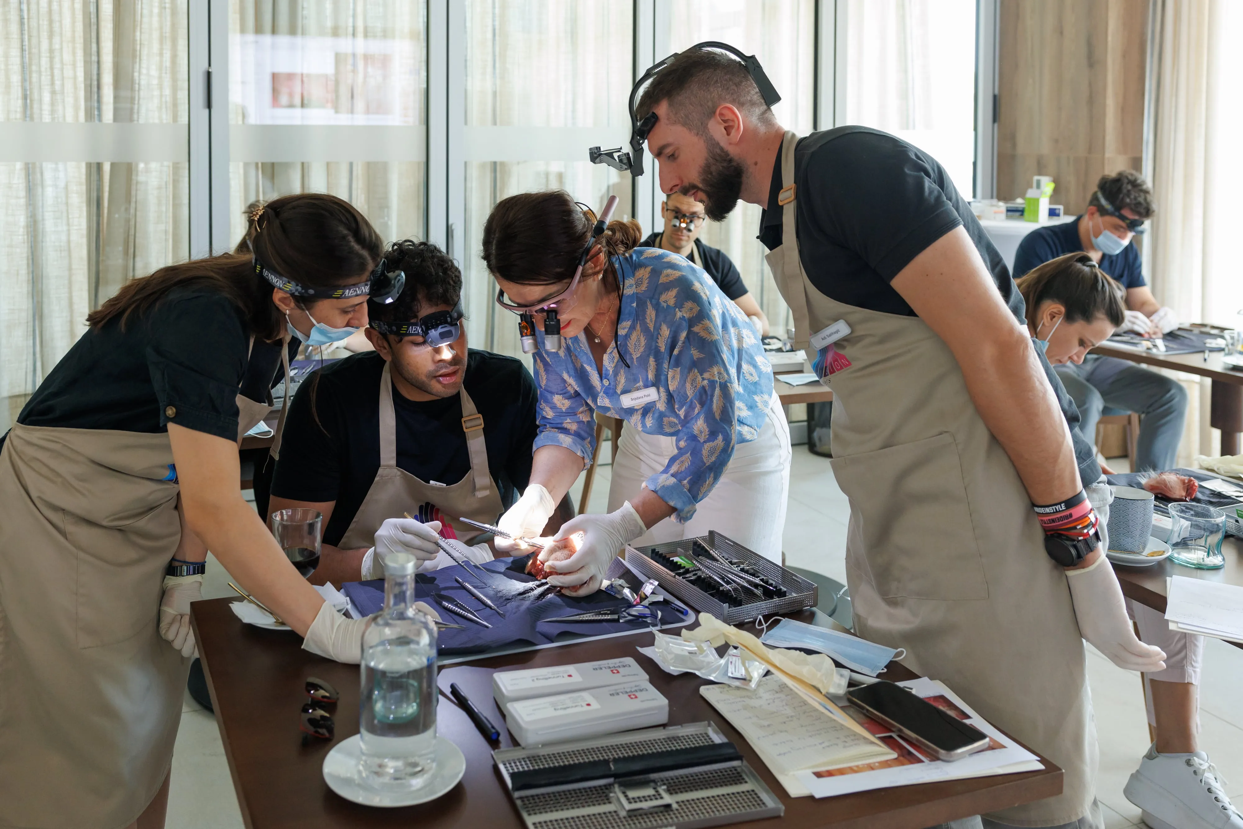 A group of people wearing aprons and magnifying headlamps closely examining an object on a table with various tools and supplies in a bright room.