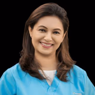 Smiling woman with shoulder-length brown hair wearing a light blue medical scrub top against a black background.