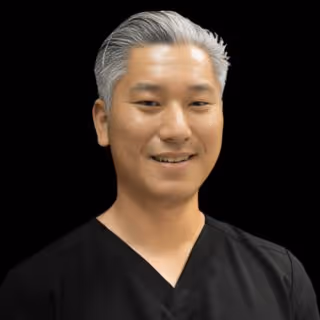 Portrait of a smiling man with gray hair wearing a black V-neck shirt against a black background.