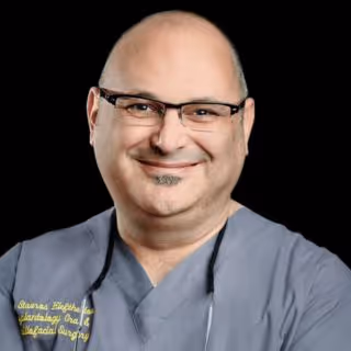 Smiling bald man wearing glasses and grey medical scrubs with a black background.