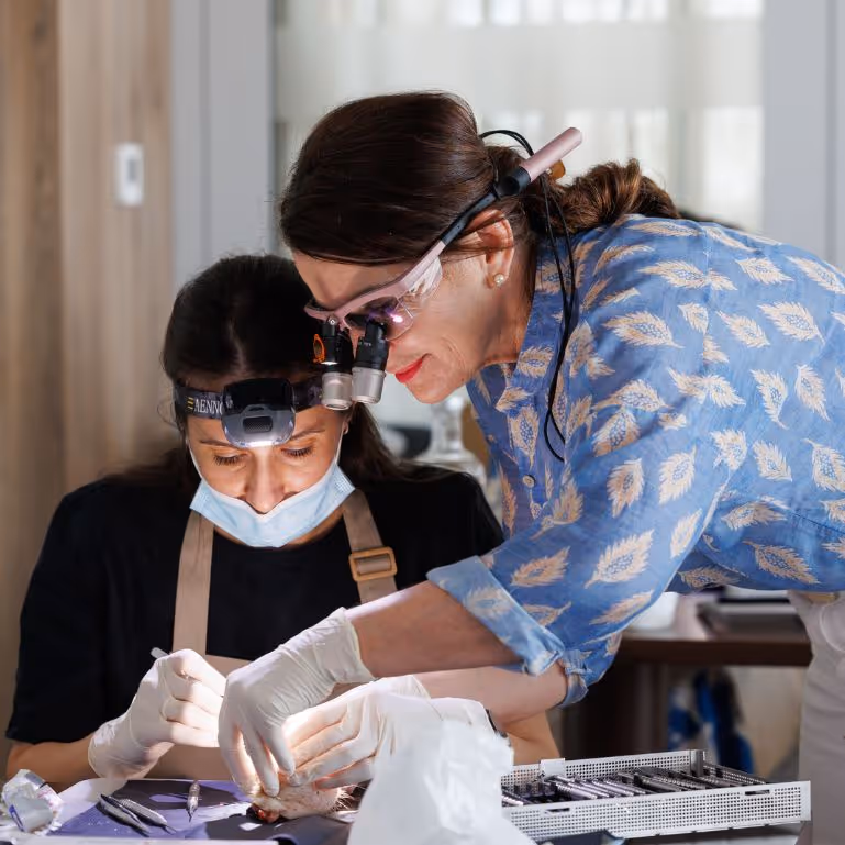 Two women wearing magnifying headlamps and gloves carefully performing a detailed task together at a workspace with tools.