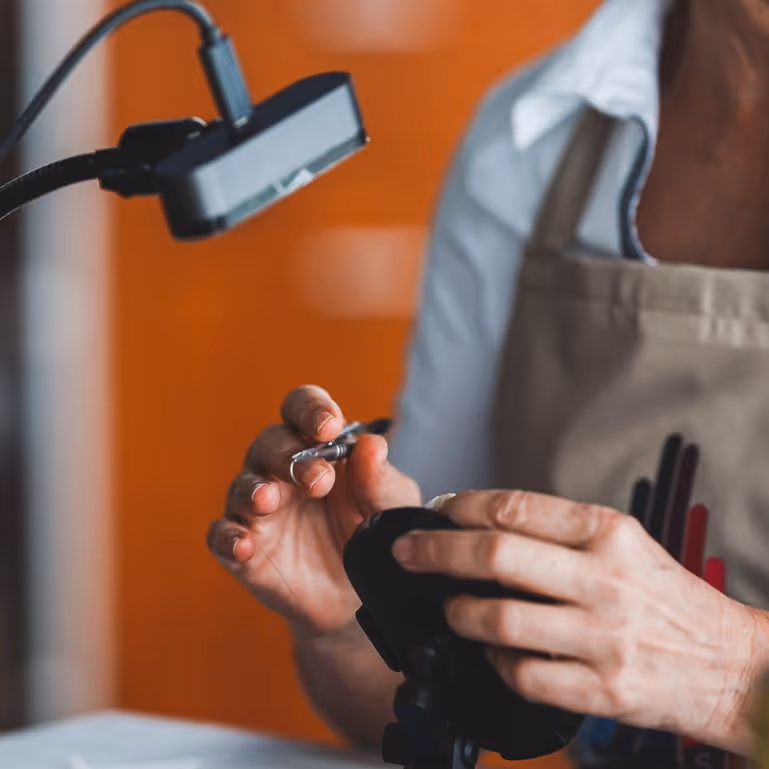Person wearing an apron holding and examining a small metallic object under a magnifying lamp.