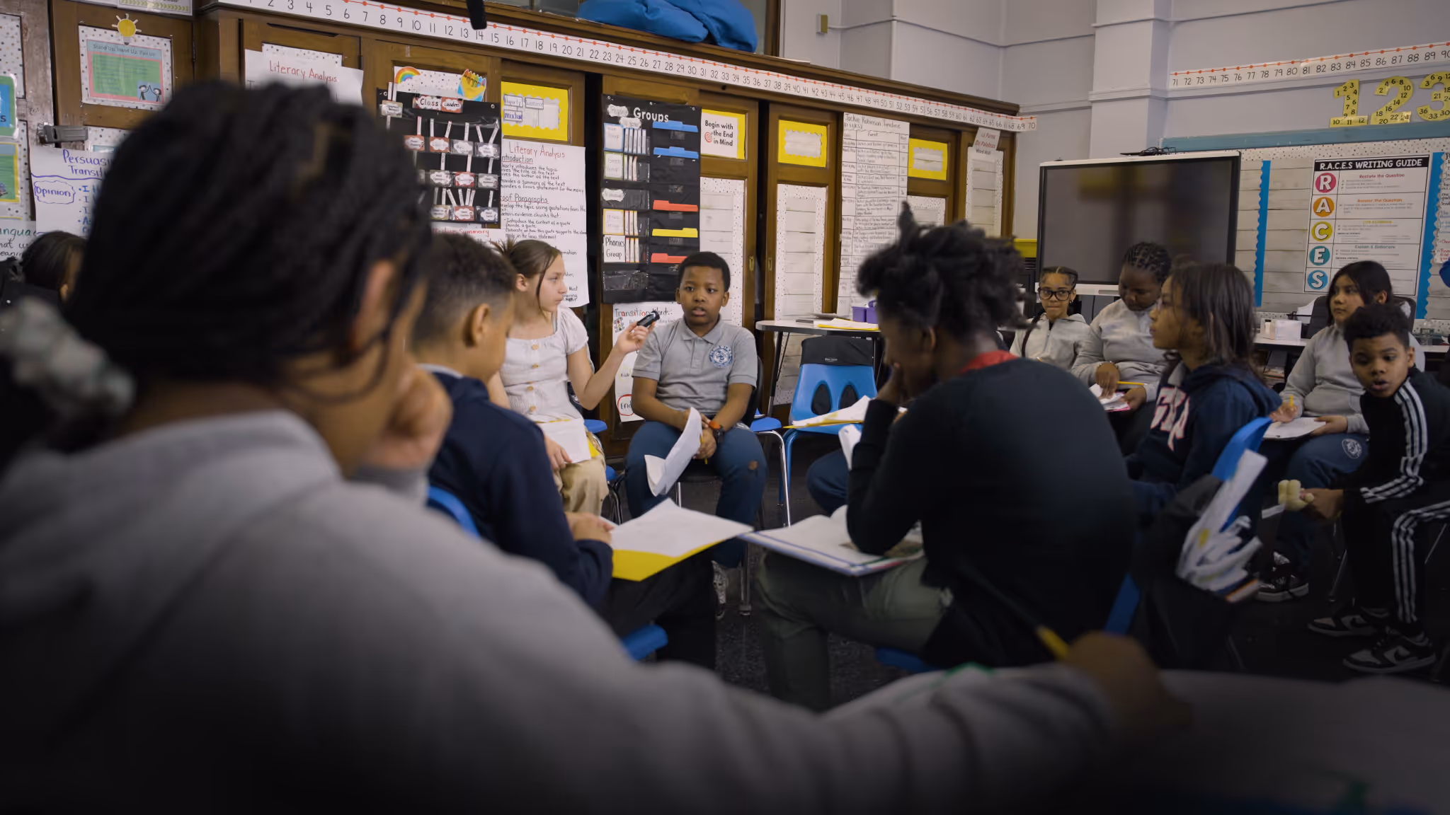 Diverse group of elementary school students sitting in a circle, engaged in a classroom discussion with notebooks and papers.