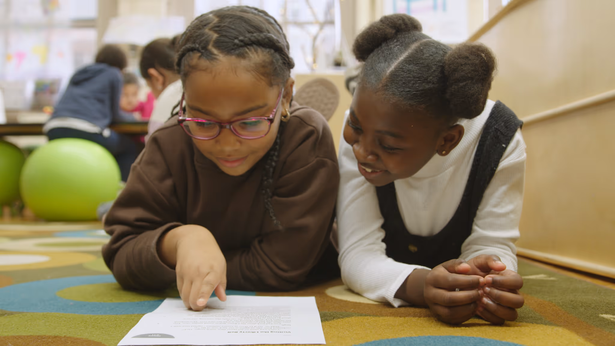 Two young girls lying on a colorful carpet, reading and discussing a document together in a classroom setting.