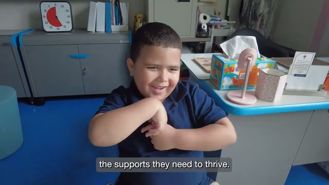 Smiling boy in a navy blue shirt sitting and using sign language in a classroom setting.