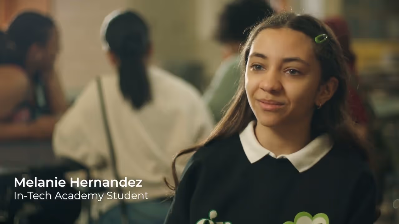 Young woman with long brown hair and a hair clip, wearing a black sweater with a white collar, smiling gently in a classroom setting with blurred students in the background.