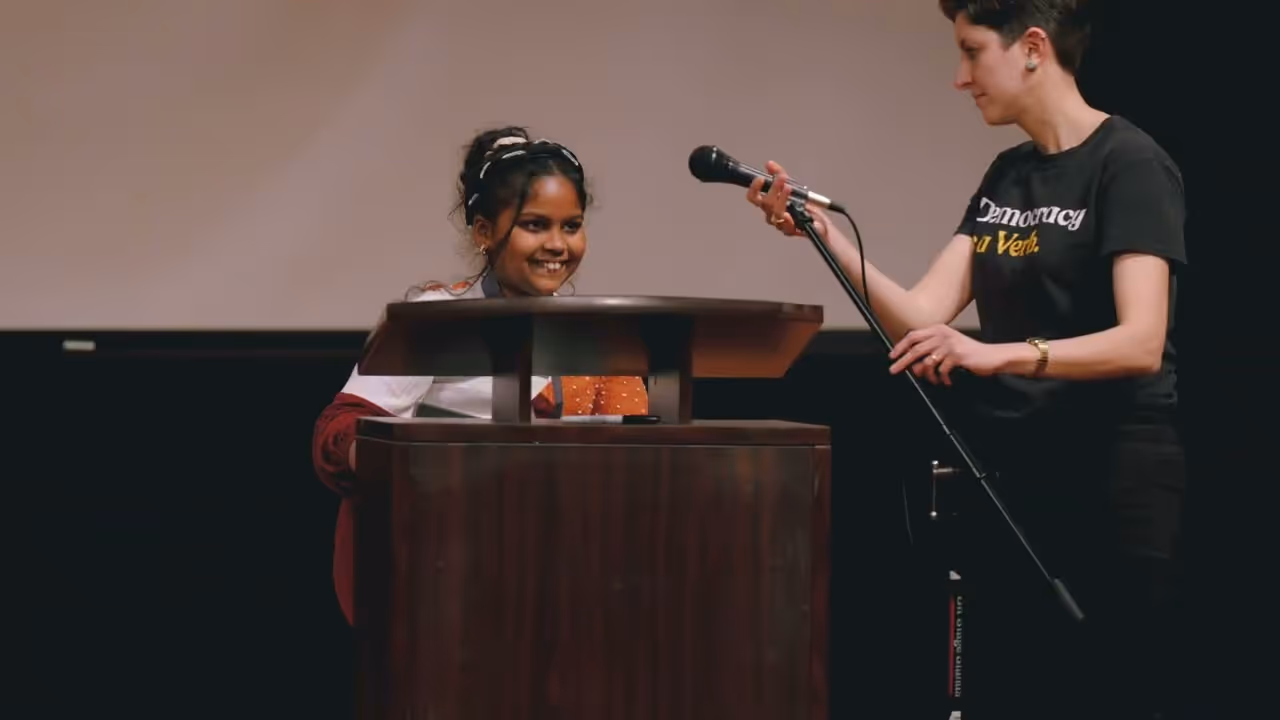 Teacher and smiling student at a classroom table with books and supplies, engaging in a learning activity.