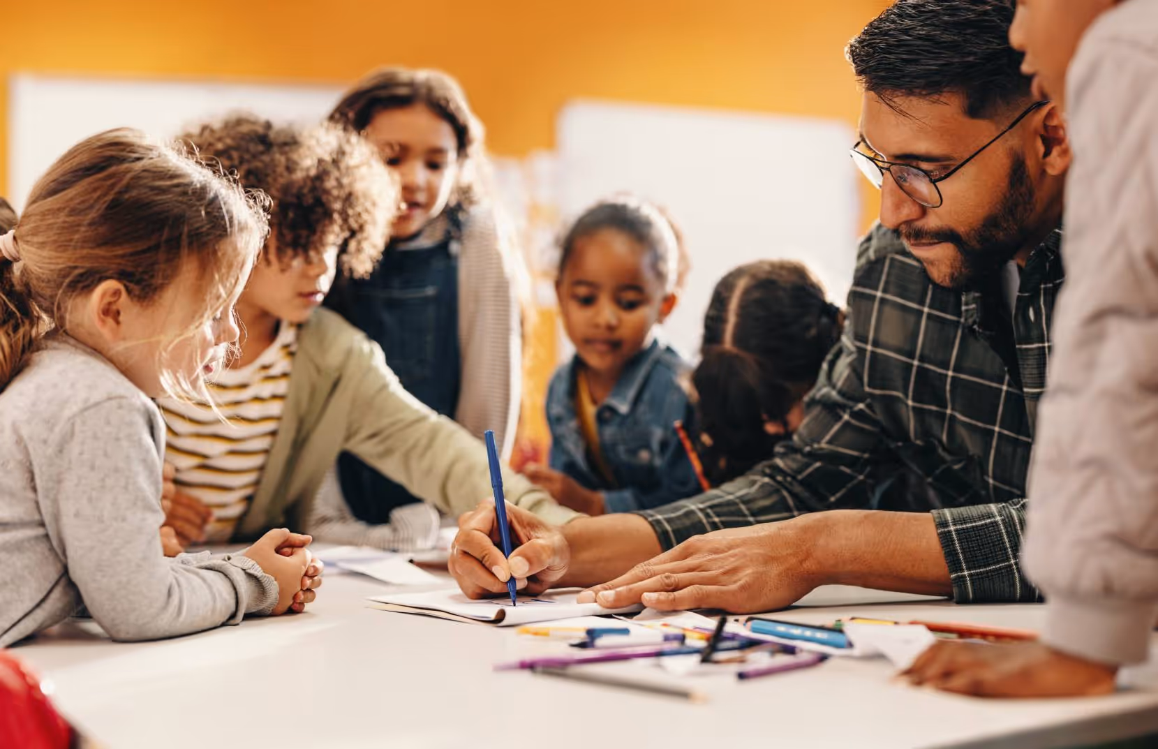 Teacher writing with a blue pen while a group of young children attentively watch around a table.
