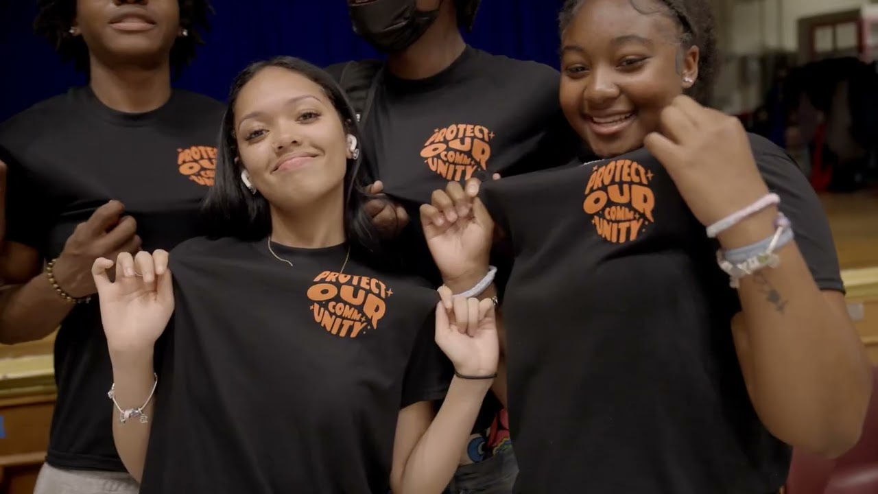Two women with braided hair smiling closely together on a couch, one gently touching the other's face.