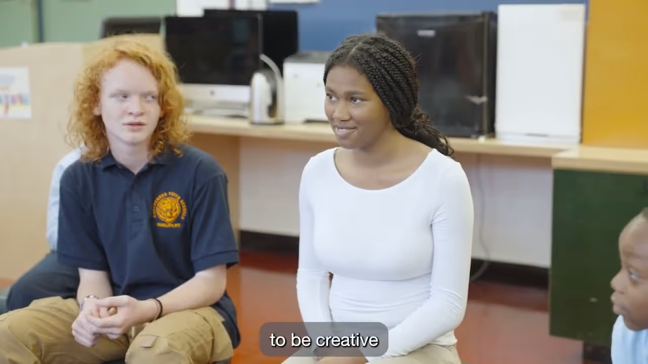Smiling man wearing glasses and a light blue blazer sitting in front of a striped wooden wall with bookshelves and framed artwork in the background.