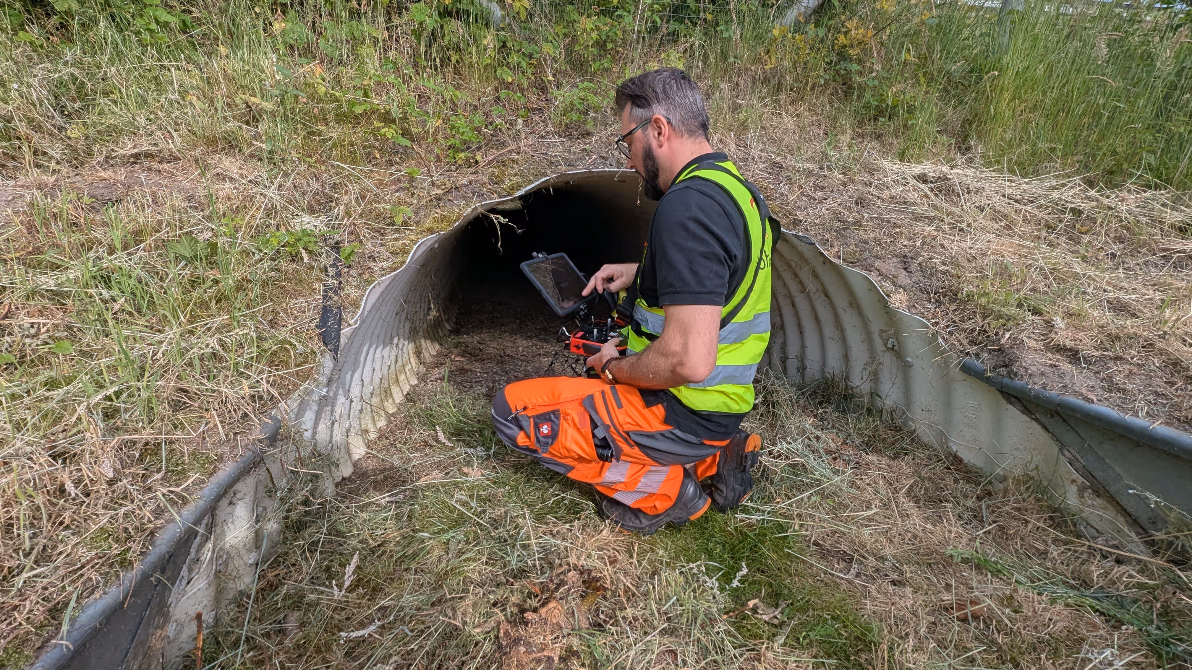 Ein Mann mit Arbeitsbegleitung hält eine Fernsteuerung für eine Drohne in der Hand. Die Drohne schwebt vor ihm. Im Hintergrund ist eine Industrieanlage zu erkennen.
