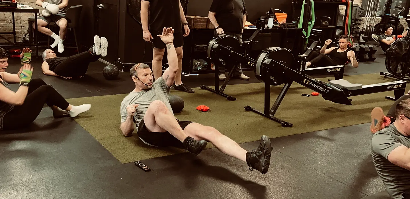 Group fitness class with participants doing floor exercises and rowing on indoor rowing machines in a gym.