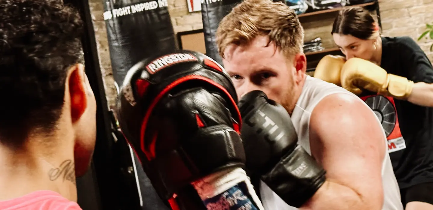 Two people sparring in a boxing gym with a trainer holding pads and a woman boxing in the background.