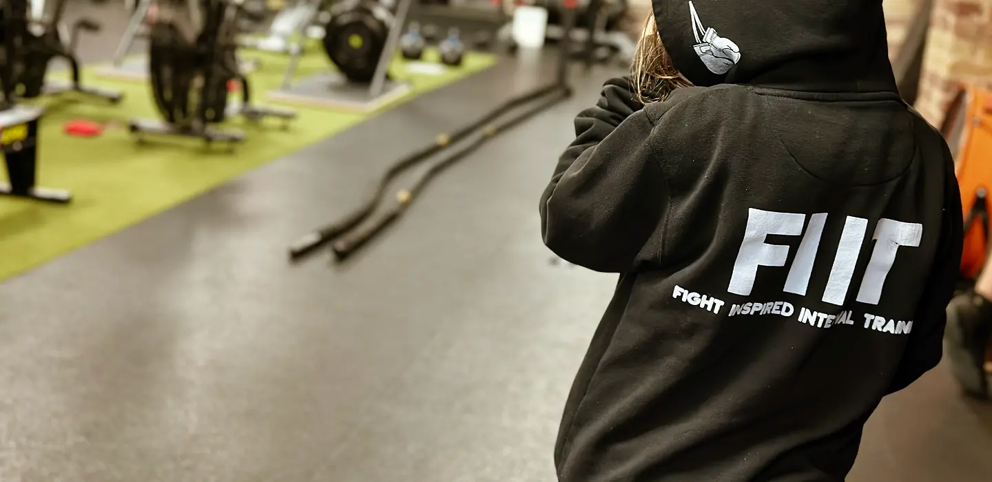 Person wearing a black hoodie with 'FIIT Fight Inspired Interval Training' text on the back, standing in a gym with workout equipment.