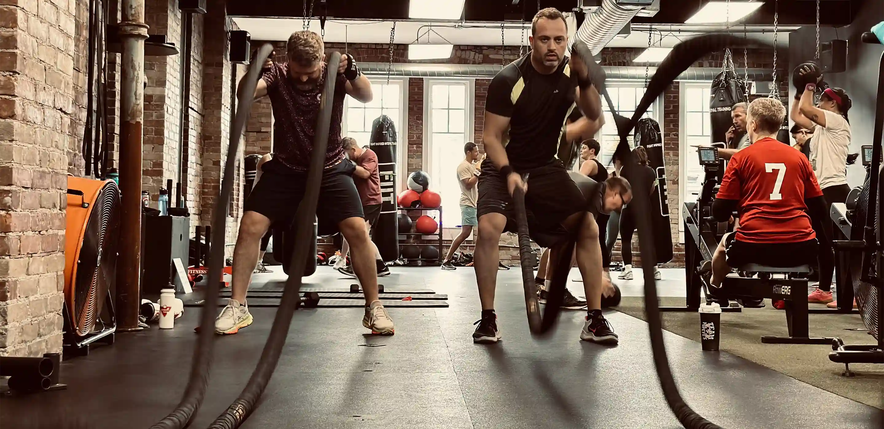 People working out with battle ropes and medicine balls in a busy gym with brick walls and large windows.
