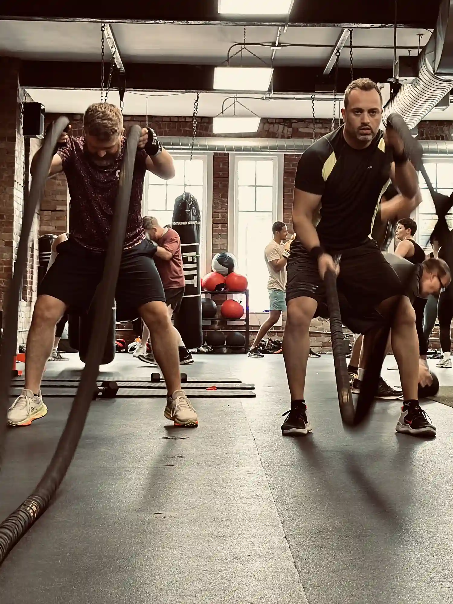 Two men intensely exercising with battle ropes in a gym with other people working out in the background.