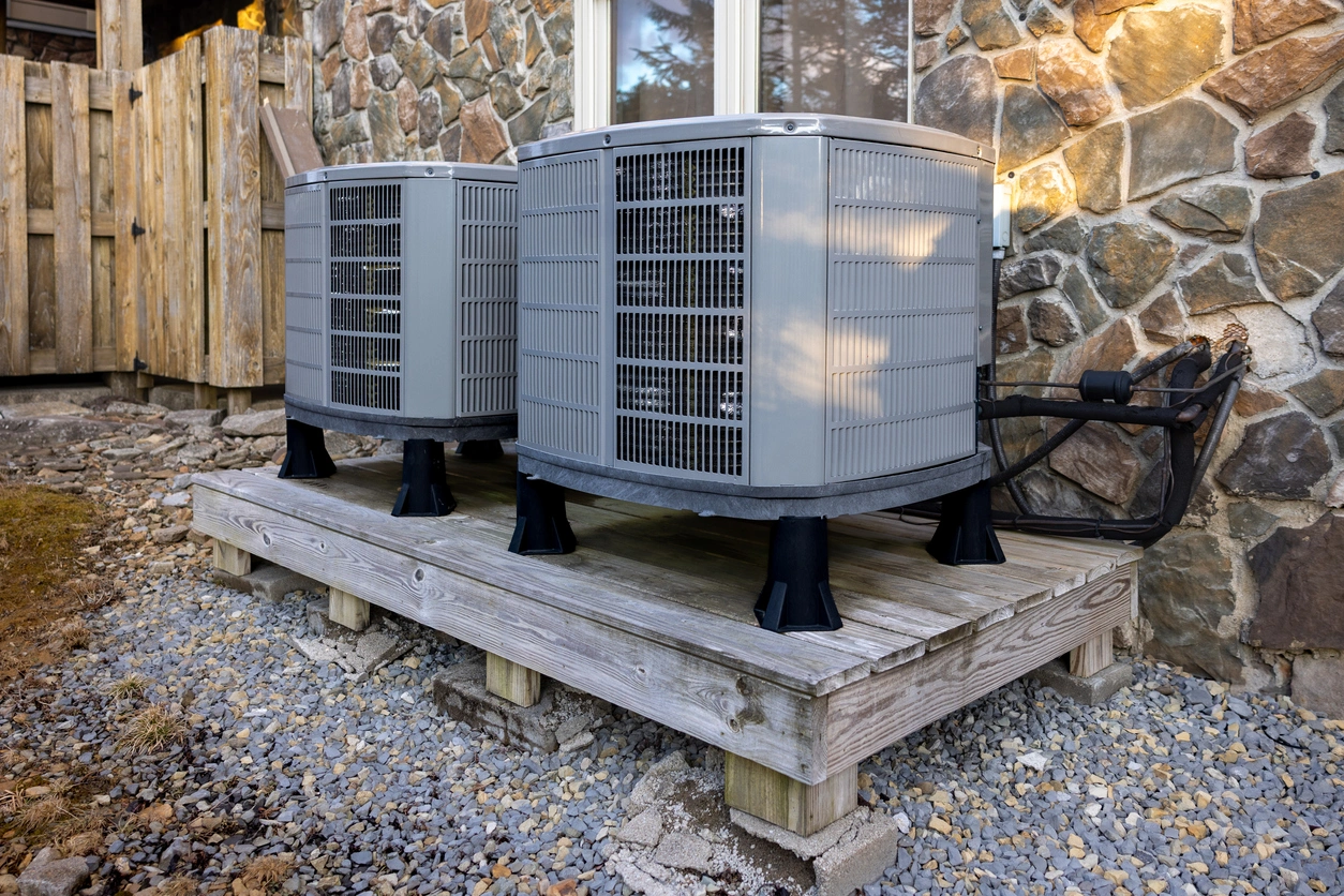Two gray air conditioning units on a wooden platform outside a stone wall building, surrounded by gravel. 