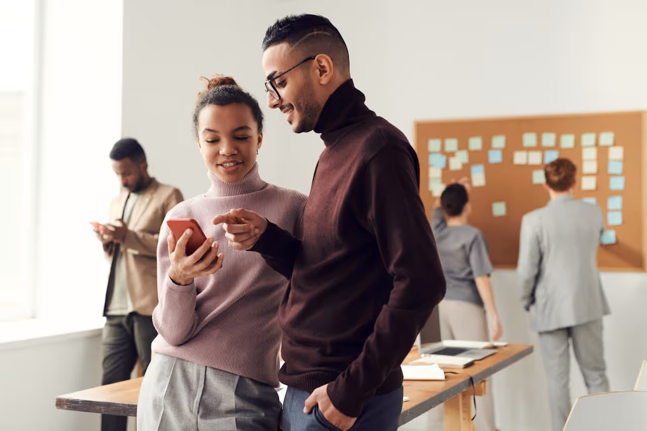 Two people in office enviroment chatting and looking at a phone.