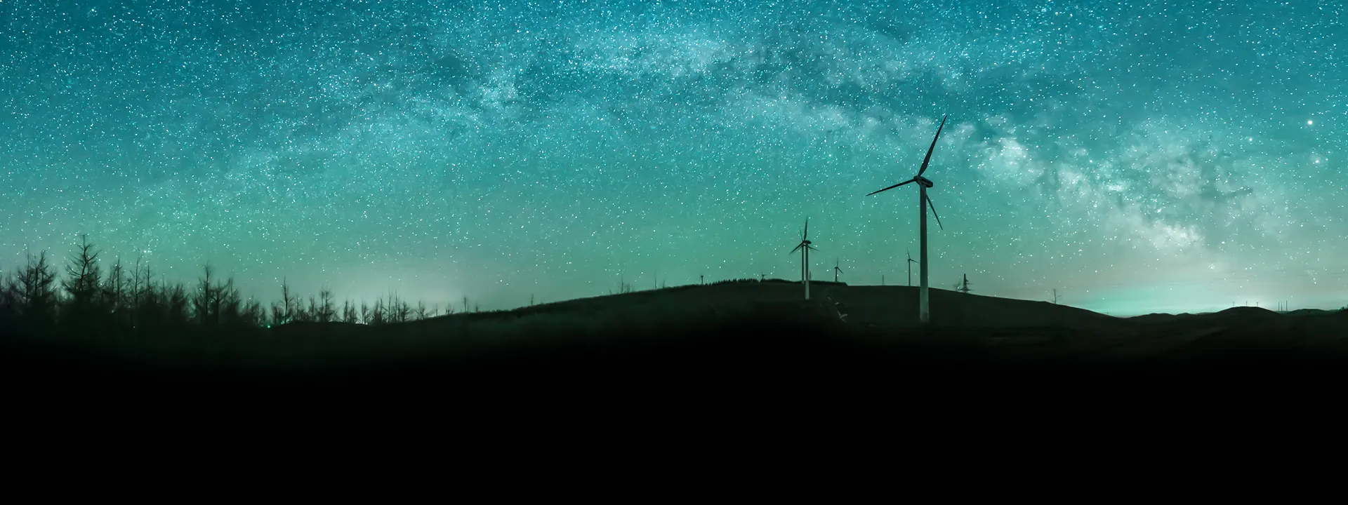 Silhouette of wind turbines on a hill under a starry, teal-colored night sky with the Milky Way visible.