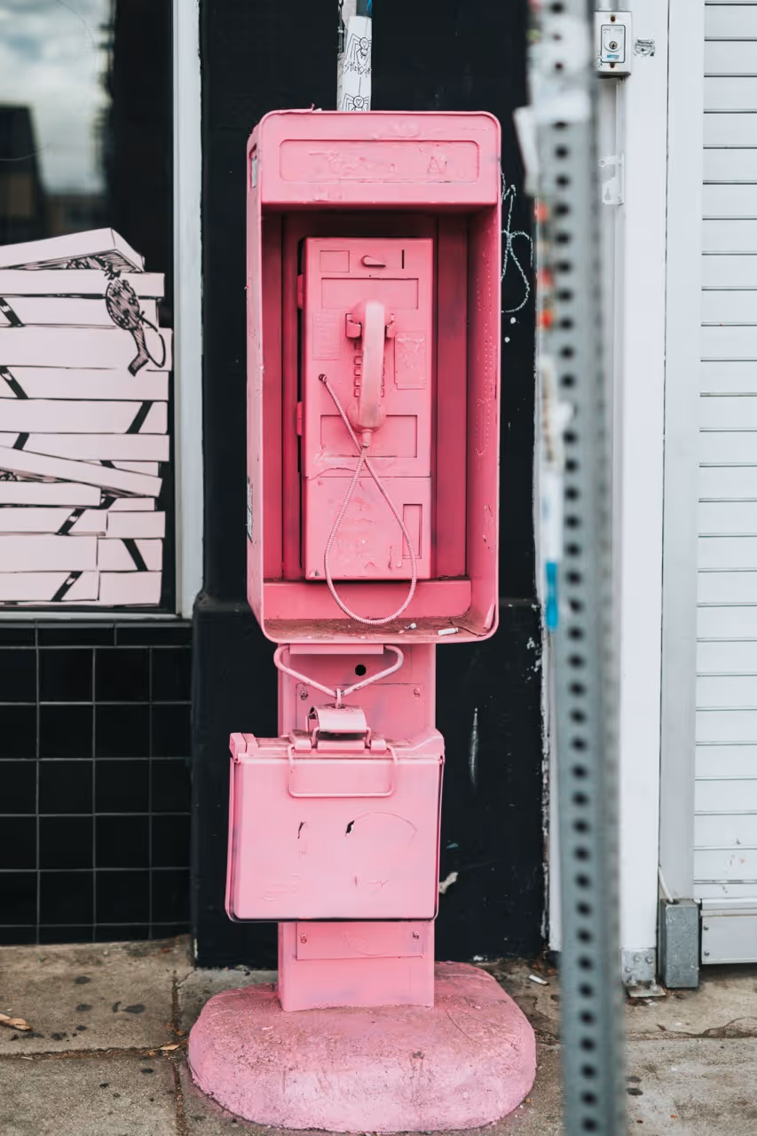 Cabina telefonica pubblica completamente verniciata di rosa con un telefono rosa all'interno, posizionata su un marciapiede urbano.
