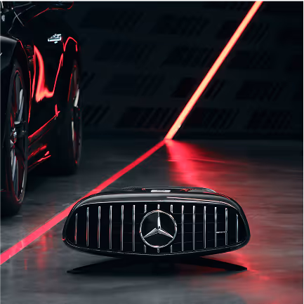 Una griglia anteriore nera con il logo Mercedes-Benz posata sul pavimento davanti a una macchina con riflessi di luce rossa.