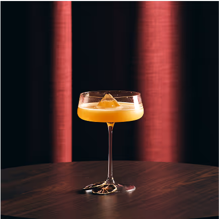 Bicchiere da cocktail con bevanda arancione e una decorazione, posato su tavolo scuro con sfondo di tende rosse sfocate.
