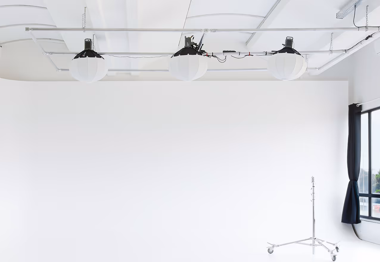 Interno di uno studio fotografico bianco con tre luci sospese al soffitto e un supporto a treppiede su ruote vicino a una finestra con tende nere.