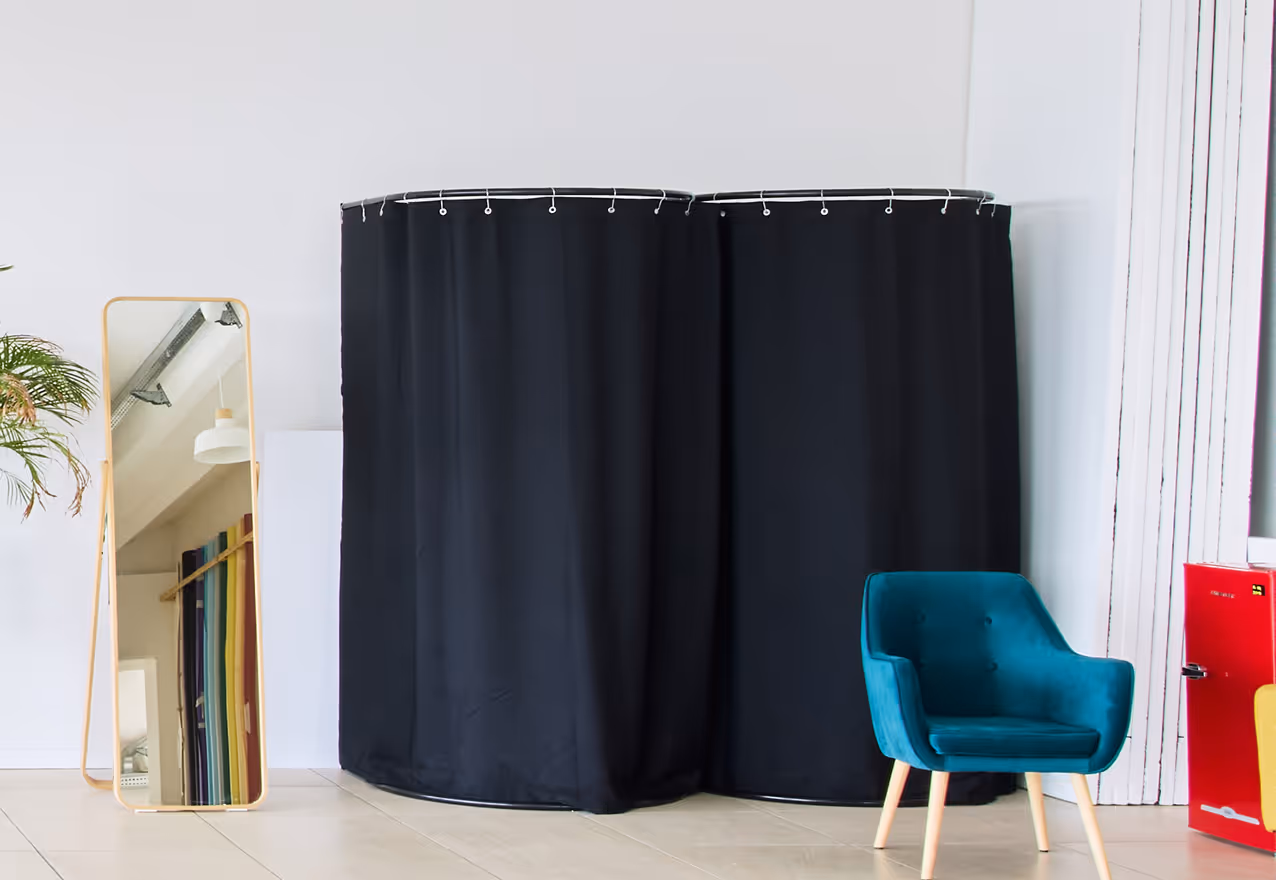 Specchio a figura intera con cornice leggera, tenda doccia nera curva e poltrona blu in una stanza luminosa con pavimento chiaro.