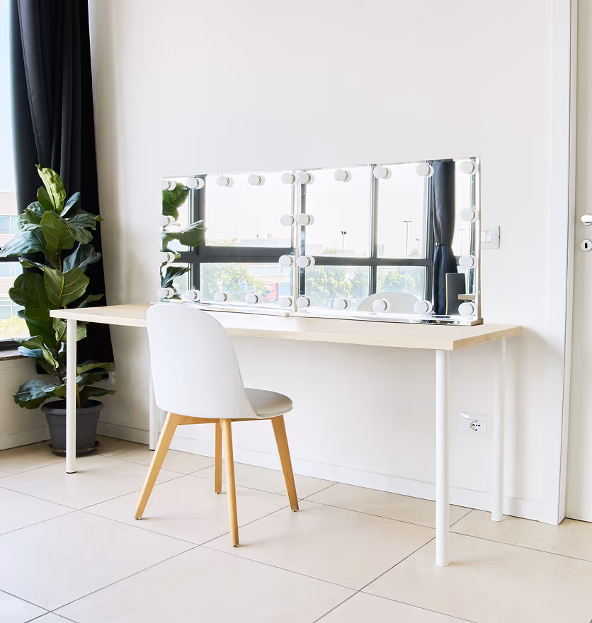 Scrivania con specchio da trucco illuminato, sedia bianca e pianta in vaso vicino alla finestra.