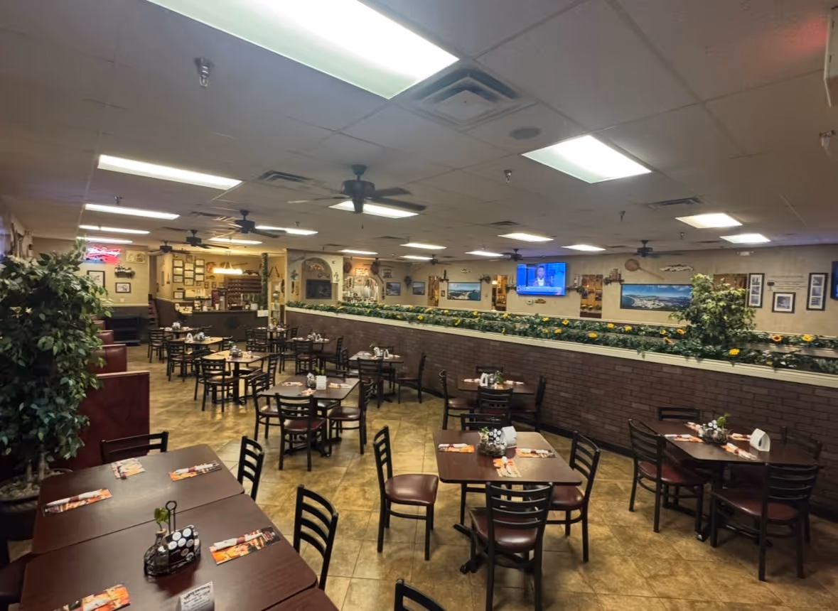 Interior of a cozy restaurant with multiple dark wood tables and chairs, ceiling fans, plants, and a TV on the wall.