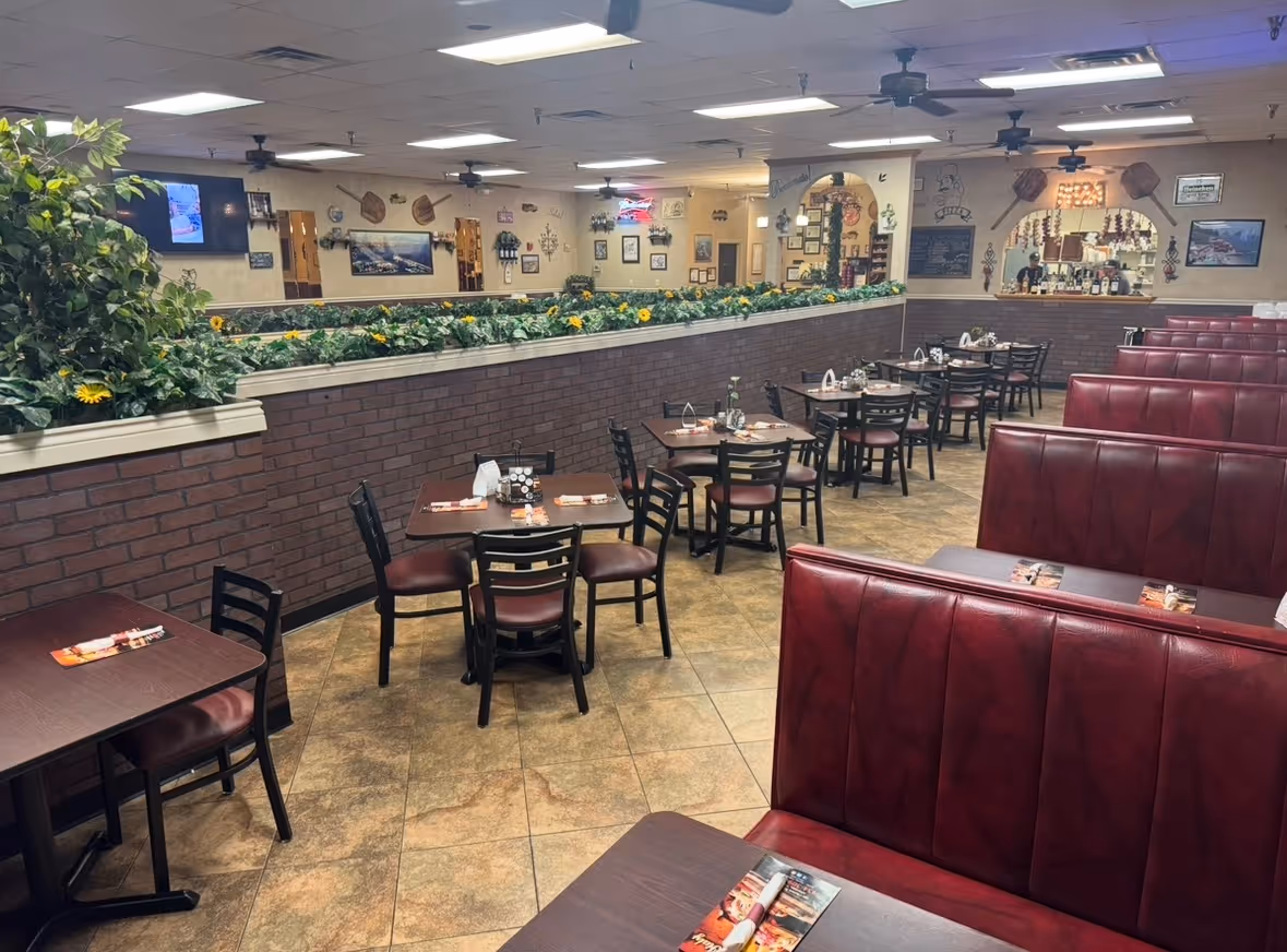 Interior of a restaurant with red leather booths, dark wood tables with chairs, beige tiled floor, and wall decorations including plants and pictures.