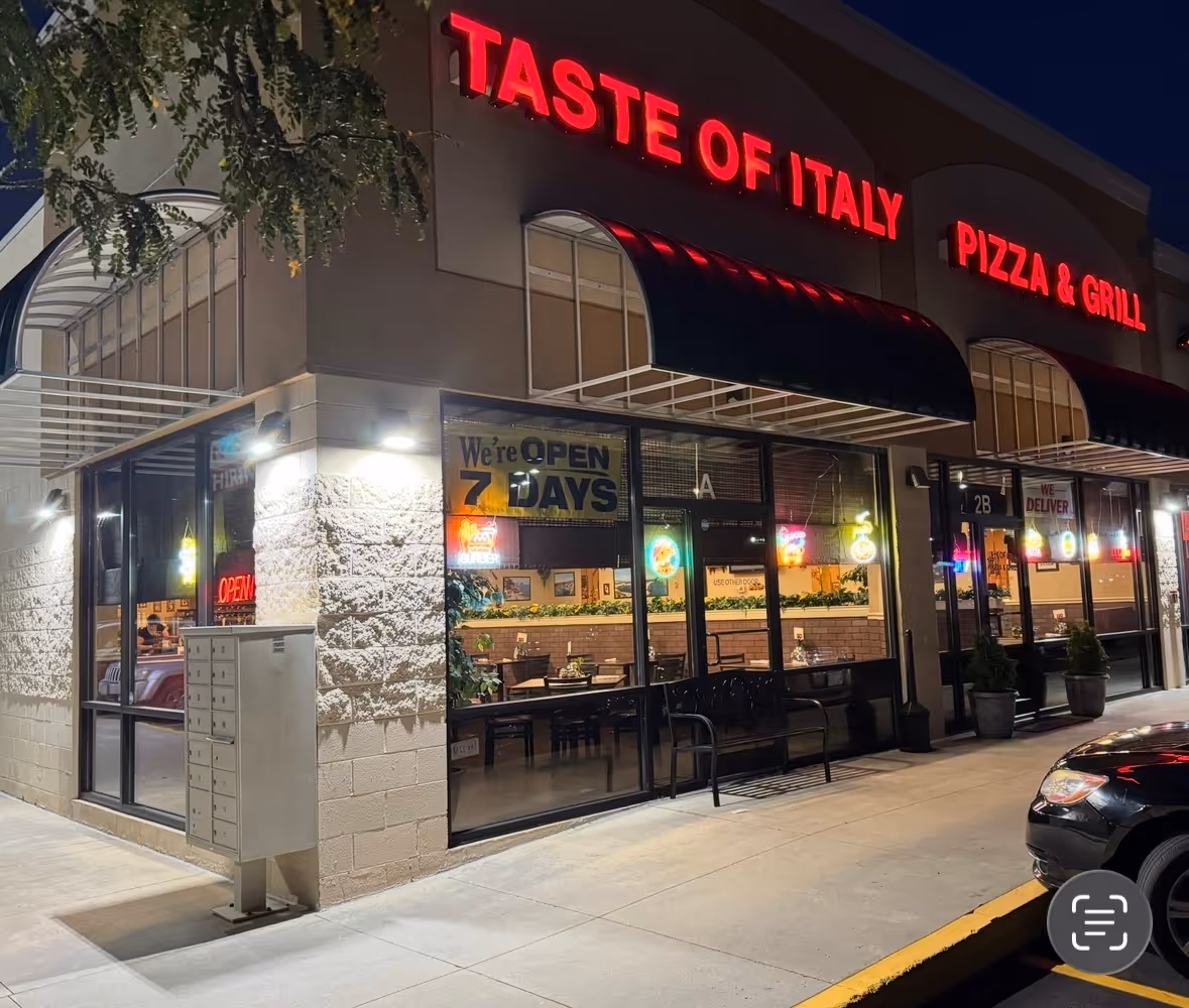Night view of the Taste of Italy Pizza & Grill restaurant storefront with illuminated red signs and visible dining area through large windows.