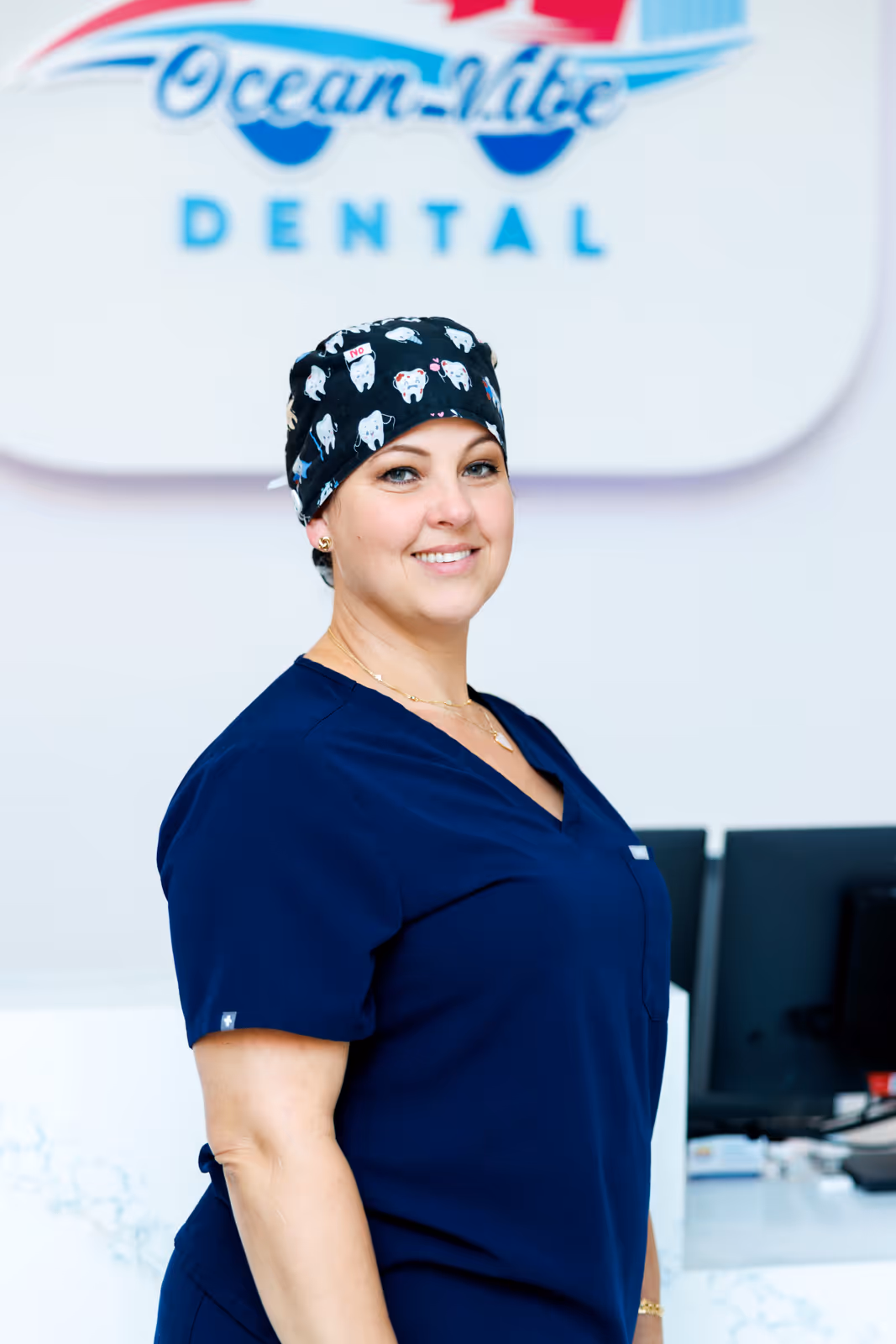 Smiling female dental professional wearing navy scrubs and a black cap with tooth illustrations, standing in a dental office.
